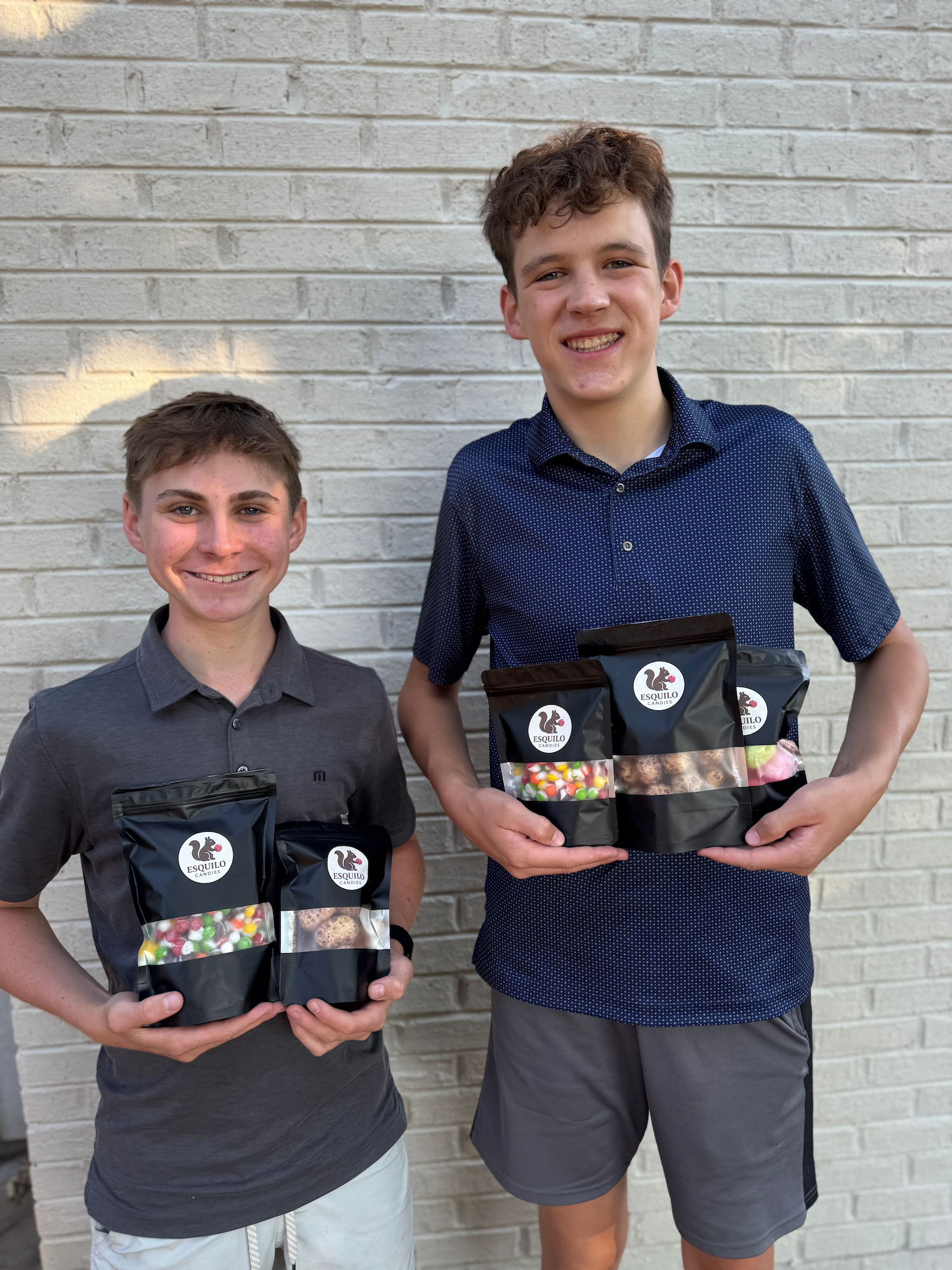 Denver Freeze Dried Candy founders Dylan + Bennett  holding bags of their candy in front of a white brick wall.